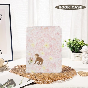 Puede incluir: Una funda de libro rosa con un estampado floral y una ilustración de mono marrón. El texto "BOOK CASE" está en la parte superior. El mono está mirando una flor. La funda está sobre una estera tejida.