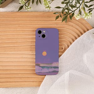 以下が含まれることがあります： 黄色い太陽と海の波が描かれた夕焼けのデザインが特徴の、ライラック色の携帯電話ケース。ケースにはカメラの切り抜きがあります。携帯電話ケースは、装飾的な緑のある木製の表面にあります。