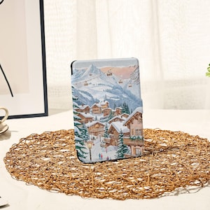 以下が含まれることがあります： 雪に覆われたシャレー、常緑樹、スキーリフトが雪をかぶった山々を背景にした冬の村のシーンが描かれたタブレットケース。ケースは黒い縁取りとテクスチャ加工された表面を持っています。