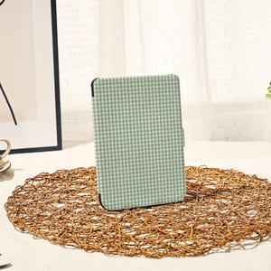 May include: A tablet case featuring a light green and white gingham pattern. The case is positioned on a circular, woven placemat. The background includes a white curtain and a framed artwork.
