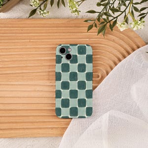 Puede incluir: Una funda de teléfono inteligente con un patrón de tablero de ajedrez verde y azul claro. La funda tiene un recorte para la cámara y se muestra sobre una superficie de madera con vegetación decorativa y tela blanca.