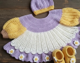 May include: A handmade crochet baby outfit in shades of lavender, white, and yellow. The set includes a dress with daisy accents, a matching bonnet, and a pair of baby shoes. The dress features yellow sleeves and a white skirt.