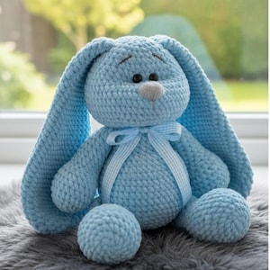 May include: A light blue crochet bunny with long floppy ears, a grey nose, and a blue ribbon bow. The soft toy has a textured appearance and is sitting on a grey surface.