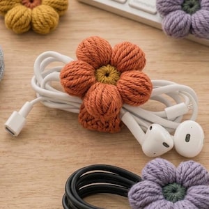 May include: A white wired earphone set coiled and secured with a crocheted orange flower cable organizer. The flower features five rounded petals and a yellow center. Additional crocheted flowers in yellow and purple are visible nearby.