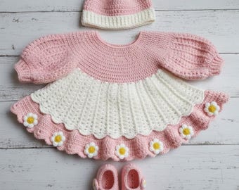May include: A handmade crochet baby outfit set in pink and white, featuring a dress, hat, and booties. The dress has a scalloped hemline adorned with daisy appliques. The hat and booties match the dress, creating a cohesive and charming ensemble.