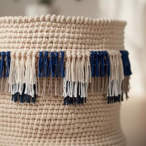 Pode incluir: Uma cesta bege, feita à mão em crochê, com uma franja decorativa. A franja apresenta secções alternadas de fio azul marinho, branco sujo e taupe. A textura da cesta é visível, mostrando os pontos apertados do trabalho de crochê. O design é simples e elegante.