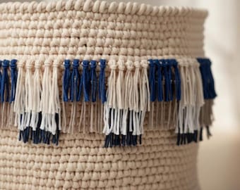 May include: A beige, hand-crocheted basket with a decorative fringe. The fringe features alternating sections of navy blue, off-white, and taupe yarn. The basket's texture is visible, showcasing the tight stitches of the crochet work. The design is simple and elegant.