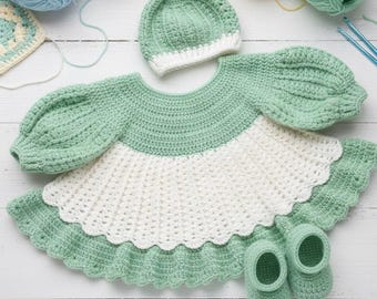 Op de afbeelding: Een handgemaakte gehaakte babyoutfit in mintgroen en wit, inclusief een jurk met geschulpte randen, een bijpassende hoed en babyslofjes. De jurk heeft pofmouwen. Garen en haaknaalden zijn zichtbaar op de achtergrond.