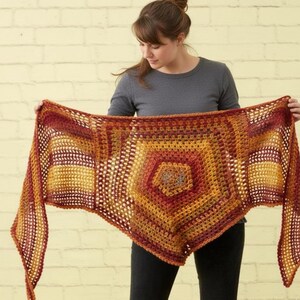 May include: A handmade, open-weave shawl in shades of orange, yellow, and red. The shawl features a geometric design with a central hexagon shape and rectangular panels. The yarn appears to be a soft, textured material, perfect for a cozy accessory.