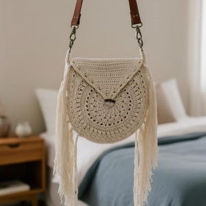 Puede incluir: Bolso bandolera de crochet color crema con diseño circular y flecos. El bolso tiene una solapa triangular y correas de cuero marrón con herrajes metálicos. El bolso cuelga frente a una cama con una manta azul.