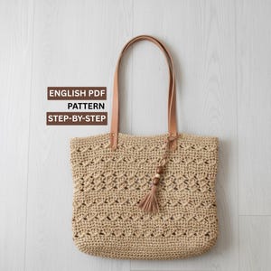 May include: A beige crochet tote bag with leather handles and a decorative tassel. The bag features a patterned design and the text "ENGLISH PDF PATTERN STEP-BY-STEP". The bag is designed for carrying items.