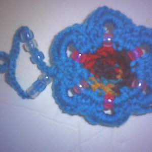 May include: A handmade, blue crocheted flower with a loop and beaded string. The flower has a multi-colored center with red, brown, and yellow hues. Pink beads are sewn into the flower's petals. A unique, handcrafted item.