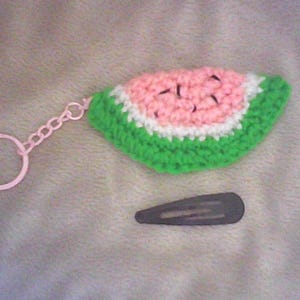 May include: A crocheted watermelon slice keychain with a pink ring and chain. The watermelon has a pink center, white rind, and green outer edge. A small, dark gray hair clip is also present.