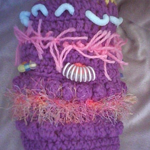 Puede incluir: Un artículo de ganchillo hecho a mano y caprichoso en tonos morado, rosa y amarillo. La pieza texturizada presenta elementos decorativos como acentos curvos blancos, hilo rosa y un adorno blanco y naranja. El artículo tiene un diseño único y lúdico.