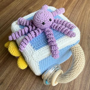 May include: A handmade crochet baby toy set featuring a lavender octopus on a blue and white striped blanket. Includes a beige rattle with wooden rings and yellow crochet accents. A soft, colorful baby gift.