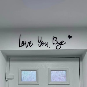Puede incluir: Texto negro en cursiva sobre una pared blanca encima de una puerta blanca que dice "Love You, Bye" con un pequeño corazón negro. La puerta tiene dos ventanas rectangulares con vidrio texturizado.