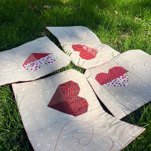 Könnte beinhalten: Mehrere handgefertigte, herzförmige, gesteppte Tischsets liegen auf grünem Gras. Jedes Tischset hat einen beigen Hintergrund mit Streifenmuster und ein rotes Herzdesign. Die Herzen sind aus verschiedenen roten Stoffen gefertigt.