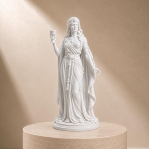 Può includere: Una statua bianca di una figura in piedi con lunghi capelli fluenti, che indossa una veste drappeggiata con vita cinta e un lungo mantello. La figura tiene un piccolo calice nella mano destra e poggia su una base circolare con pieghe.