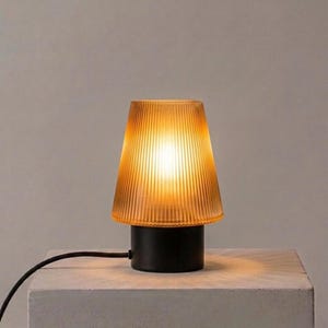 Puede incluir: Una pequeña lámpara de mesa ámbar con una pantalla de vidrio acanalado y una base negra. La lámpara está encendida, emitiendo un brillo cálido. El cable es negro.