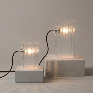 Peut inclure: Deux lampes en verre transparent avec des détails côtelés verticaux, chacune éclairée d'une lueur chaude. Les lampes sont reliées par des cordons noirs et reposent sur des socles carrés en béton gris clair. Un élément de décoration intérieure moderne.