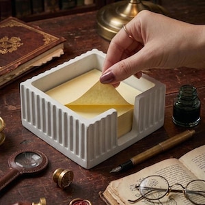 May include: A white concrete memo pad holder with vertical grooves, filled with yellow sticky notes. A hand is pulling a note from the pad. Other items on the wooden surface include a magnifying glass, ink bottle, and a vintage book.