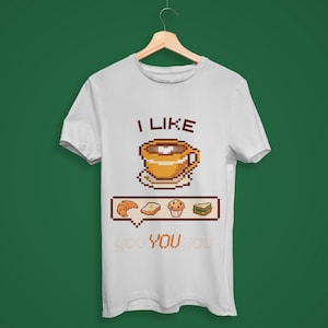 Op de afbeelding: Wit t-shirt met een pixelontwerp met een koffiekopje, croissant, brood, muffin en geld. De tekst "I LIKE YOU" staat boven en onder de afbeelding. Het shirt hangt aan een houten hanger.