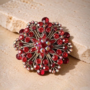 May include: A circular brooch featuring a dark gold-toned metal filigree base adorned with numerous round red rhinestones and a central cluster of iridescent, multi-colored crystals arranged in a symmetrical, floral-inspired pattern.