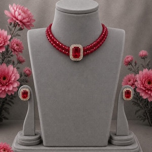 May include: A double-strand necklace featuring small red beads and a central rectangular red pendant framed by clear stones, accompanied by matching rectangular red stud earrings with clear stone borders, all displayed on a gray velvet stand.