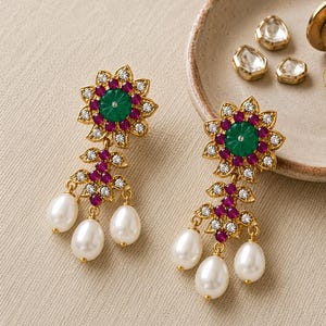 May include: A pair of gold-tone drop earrings featuring a central green carved stone surrounded by small red stones and clear crystals in a floral arrangement. Below the flower, a gold leaf-shaped structure holds three white teardrop pearls.