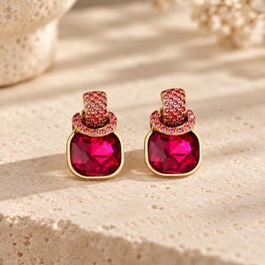 May include: A pair of gold-toned earrings featuring a large, square-cut, deep pink crystal drop suspended from a circular hoop. The hoop and the top attachment are encrusted with numerous small, sparkling pink stones, creating a vibrant contrast.