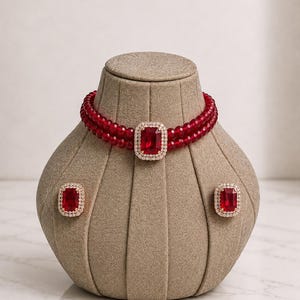 May include: A double-strand red beaded choker necklace featuring a central rectangular red stone surrounded by a halo of small clear stones, accompanied by a pair of matching rectangular red stone stud earrings with clear stone halos.