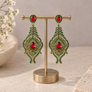 May include: A pair of gold-toned chandelier earrings featuring a teardrop-shaped red center stone surrounded by small green crystals. The earrings have a decorative filigree-style frame and a smaller matching red teardrop stone at the top post.