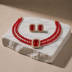May include: A jewelry set consisting of a double-strand necklace and matching stud earrings. The necklace features small red faceted beads and a central rectangular red stone pendant framed by clear crystals. The earrings match the central pendant.