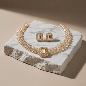 May include: A double-strand necklace and matching stud earrings set featuring faceted champagne-colored crystals. The necklace includes a central rectangular pendant with a crystal-encrusted border, while the earrings mirror this rectangular design.