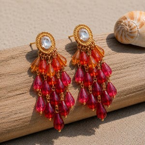 May include: A pair of gold-tone earrings featuring a circular stud with a central clear faceted stone. Cascading from each stud are multiple strands of teardrop-shaped beads in gradient shades of orange, bright red, and deep crimson red.