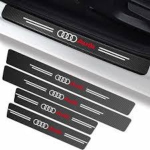 May include: Black carbon fibre car door sill protectors featuring the Audi logo in white and red. These protectors have a textured surface and are designed to prevent scratches. The set includes multiple pieces for different door sizes.