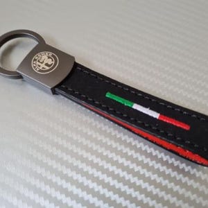 以下が含まれることがあります： Alfa Romeoのロゴが入った金属製のプレートと金属製のリングが付いた黒い革製キーホルダー。キーホルダーは赤、白、緑のストライプのデザインで、端に赤いアクセントが付いています。