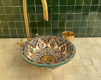 May include: A decorative ceramic sink with a colorful, intricate pattern. The sink has a brass faucet and a small brass cup next to it. The sink is set against a green tiled wall and a light-colored tiled surface.
