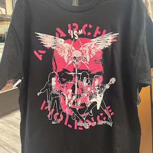 以下が含まれることがあります： ピンク色のバンドのグラフィック、頭蓋骨と翼、そして「ARCH IS HOLY」と「PUNKS IS HIPPIES」という言葉が特徴の黒いTシャツ。 Tシャツはラックに吊るされています。