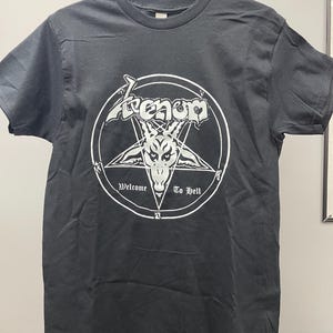 Può includere: T-shirt nera con grafica bianca raffigurante un pentagramma, una testa di capra e il nome della band "Venom". Il testo "Welcome To Hell" è incluso. La maglietta è appesa a una gruccia nera.