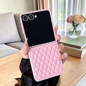 Può includere: Una custodia per telefono rosa trapuntata con schermo nero e ritagli per la fotocamera. La custodia è tenuta aperta, rivelando lo schermo del telefono. La custodia ha un motivo a rombi e una finitura liscia e opaca.