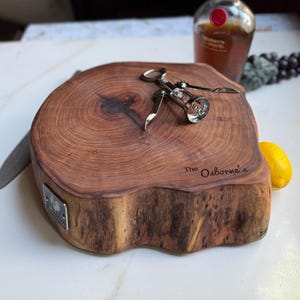May include: A round, wooden cutting board with a natural edge, featuring a silver corkscrew. The board has visible wood grain and the inscription "The Osborne's". A bottle of Maker's Mark bourbon is in the background, along with a lemon.
