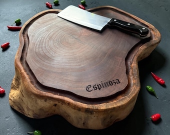May include: A thick, natural-edge wooden cutting board with a dark brown finish and a visible grain pattern. The board features the engraved name Espinoza and a deep juice groove. A silver-colored metal cleaver with a black handle rests on top.