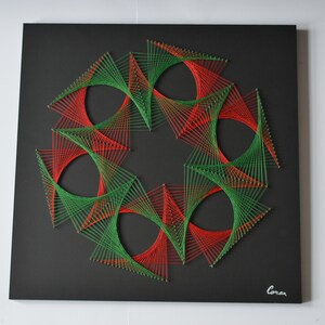 May include: A geometric string art piece on a black square canvas. The design features a circular pattern of red and green threads, creating a vibrant and symmetrical artwork. The artist's signature is in the bottom right corner.