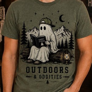 May include: An olive green heathered t-shirt featuring a graphic of a ghost wearing a baseball cap and reading a book titled Ghost Stories while sitting on a log in a forest. The text Outdoors & Oddities is printed below the illustration.