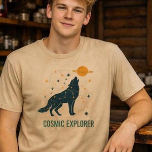 May include: Tan short-sleeved t-shirt featuring a dark blue wolf howling at a yellow planet with rings. The design includes constellations and the text "COSMIC EXPLORER" in green.