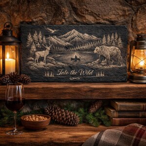 May include: A rectangular black slate sign with an engraved wilderness scene. The design features a lake, mountains, a deer, a bear, and the text "Into the Wild". The sign is displayed on a wooden shelf with lit lanterns and a glass of wine.