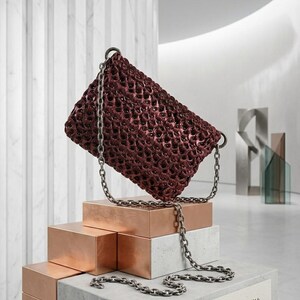 May include: A burgundy woven handbag with a chain strap rests on a display of copper and concrete blocks. The bag has a unique open weave design. The display includes a sign that reads "THE WOVEN FORM: A STUDY IN TEXTURE."