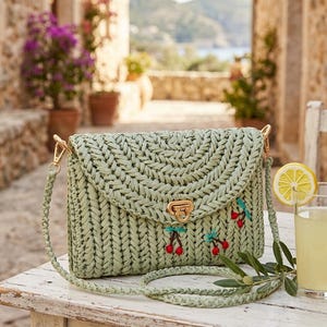 Puede incluir: Un bolso tejido verde claro con cierre de solapa y cierre dorado. El bolso presenta cerezas bordadas y una correa para el hombro a juego. El bolso está sobre una mesa de madera blanca.