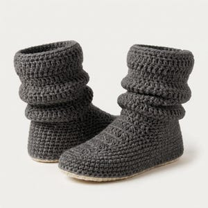 May include: A pair of dark grey ankle-high boots crafted from thick, textured crochet yarn. The boots feature a slouchy, folded cuff design around the ankle and a solid, rounded toe construction with a flat, light-coloured sole base.